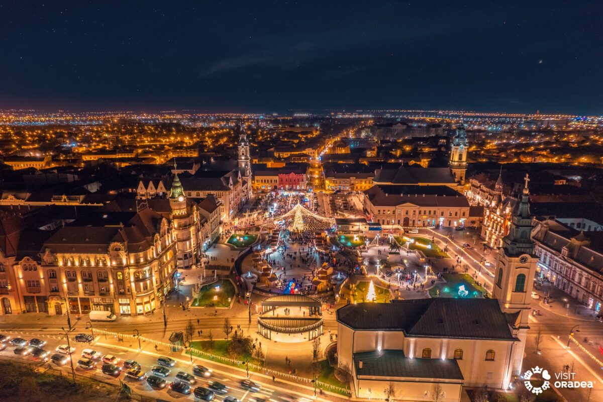 Târgul de Crăciun din Oradea - Piața Unirii văzută de sus noaptea, cu palatele Art Nouveau luminate, bradul festiv și cupola de luminițe