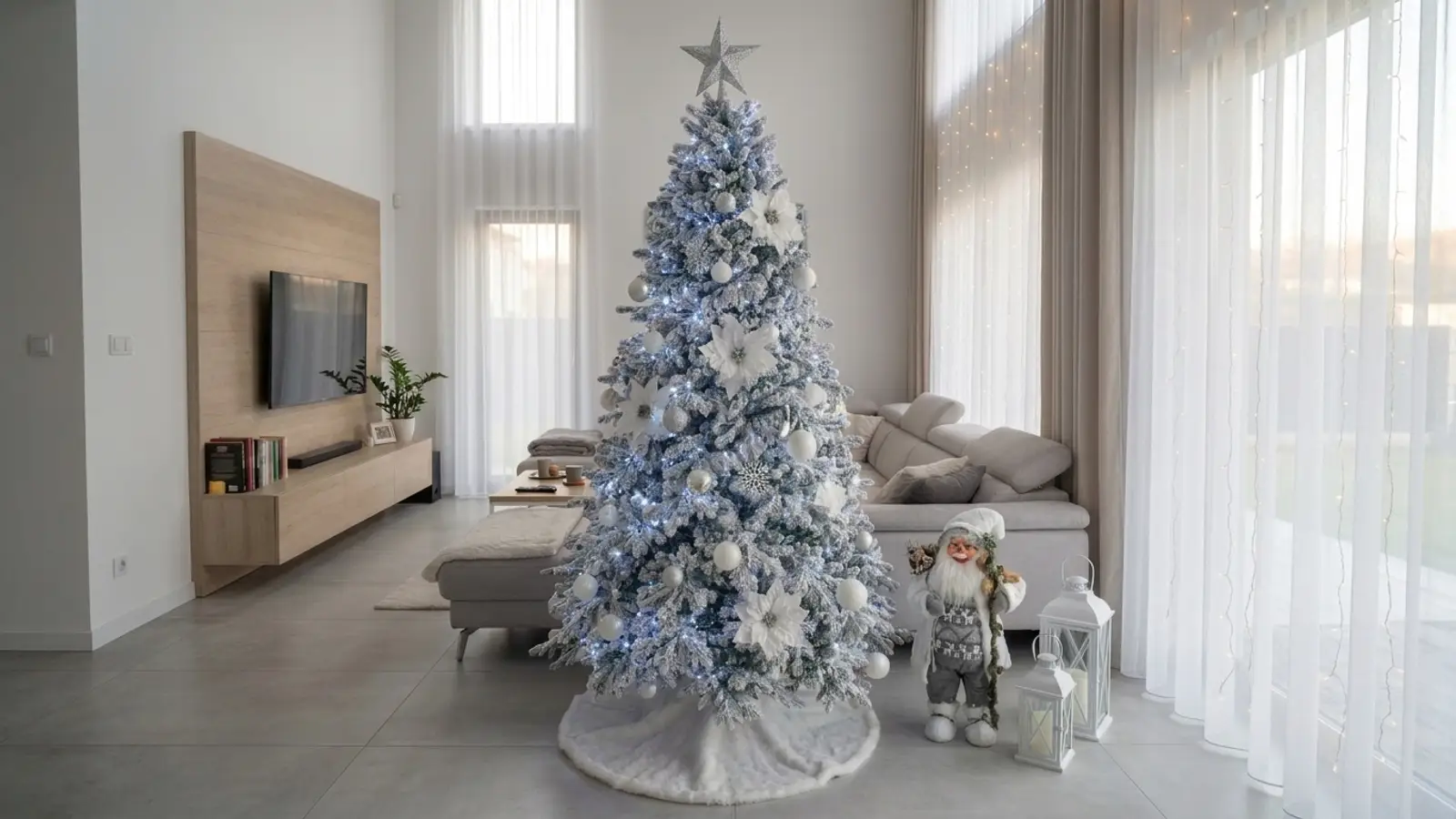 Brad artificial nins Imperial Snow de 230 cm, împodobit cu globuri albe și argintii, flori albe și lumini reci, cu o figurină de Moș Crăciun și felinare albe alături, într-un living cu gresie gri