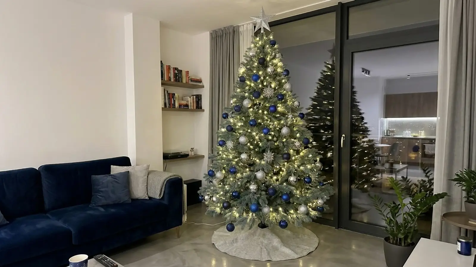 Brad artificial verde-albăstrui Imperial Grand Sapphire de 250 cm, împodobit cu globuri albastru închis și argintii și fulgi de nea, lângă o canapea bleumarin, seara, într-un apartament cu pardoseală din microciment
