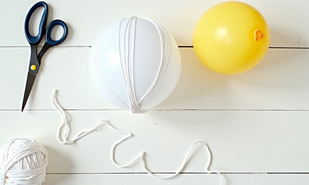 Balon umflat înfășurat în sfoară albă, lângă un ghem de sfoară și o foarfecă, pentru un glob de Crăciun făcut în casă